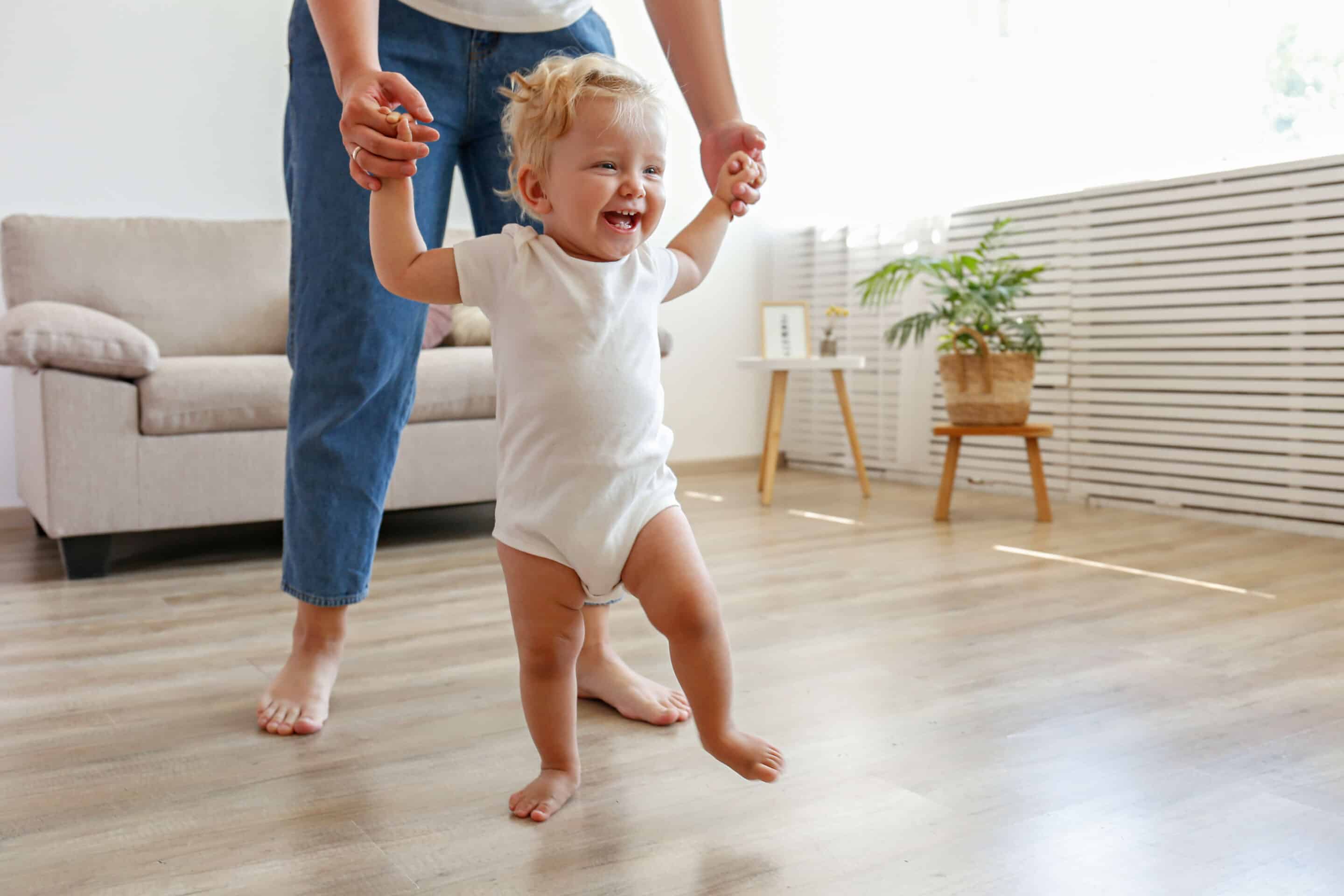 First steps of a little girl. A blond toddler learning to walk at home with the help of loving mother.